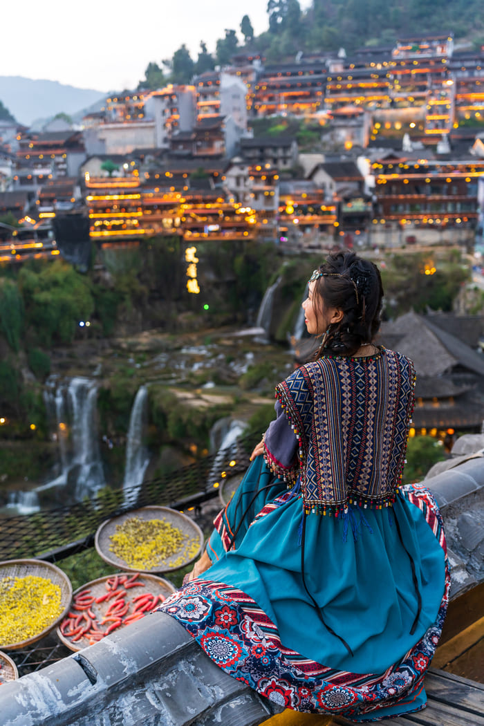 Mujer vestida con un colorido traje tradicional sentada de espaldas en un tejado, contemplando un antiguo pueblo chino iluminado y sus cascadas al atardecer.